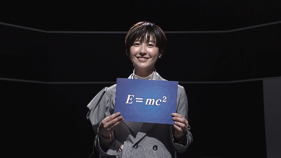 スタジオ風景写真ワンショット。女性がE=mc2と書かれたフリップを持っている