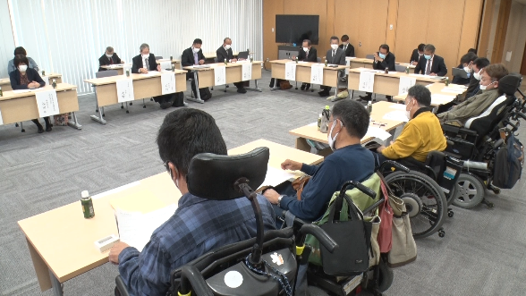 番組のスクリーンショット。会議室で会議が行われており、スーツ姿の人や車椅子に乗っている人もいる